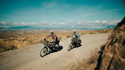 Two people ride loaded bicycles along a dusty track through mountainous terrain.