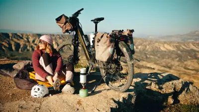 A person sits on a rock beside a loaded bike and camping gear overlooking a wide mountain landscape.