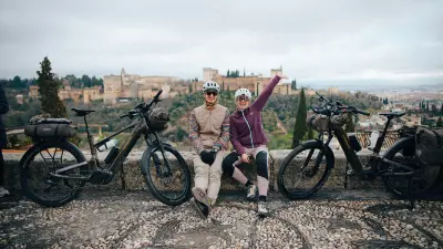 Two cyclists resting with touring bikes on a viewpoint above a historic city.