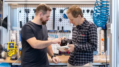 Two people examining an e bike motor component in a workshop with tools on the wall.