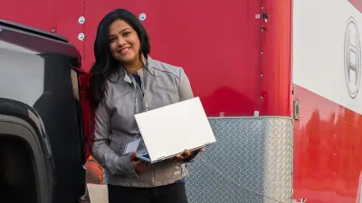 Das Bild zeigt Bosch Mitarbeiterin Bhavana. Sie hat einen Laptop in der Hand, steht vor einem großen Fahrzeug und lächelt in die Kamera.
