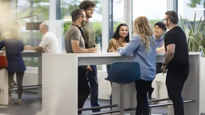 6 employees, including women and men, are seen at a workplace. They are facing each other and are in a conversation, smiling at each other. In the background there is an office environment with a large window front, plants and other employees.