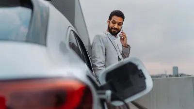 A man with a mobile phone standing next to his car. In the foreground you can see that the car is currently fueled.