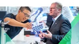 Im Vordergrund steht Stefan Hartung, während im Hintergrund ein Mitarbeiter und ein Roboterarm zu sehen sind. 