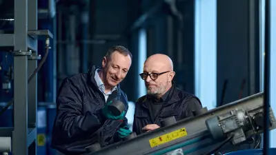 Two men in factory, one bearded and with glasses. The other man holds a fist-sized metallic object in his hand which the two men observe.