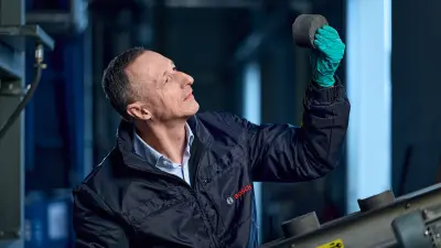Mann in Fabrikhalle hält faustgroßen metallischen Gegenstand in der Hand nach oben und blickt hinauf zu dem Objekt.