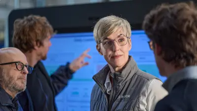 In front of a large monitor, a woman with short blonde hair and glasses is in conversation with several people.