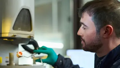 Man with full beard sits at his desk, holding a small object in a pair of pincers.