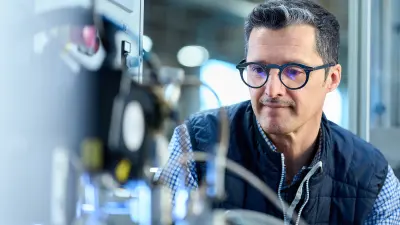 Smiling into the camera, a man leans forward to look at a machine with cables and wires.