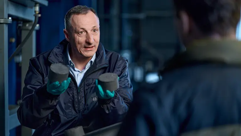 Man in a factory holds two fist-sized metallic objects in his hands.