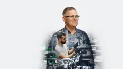 A man stands in a factory in front of a workbench and inspects individual parts of a steering system.