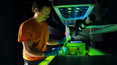 A man wearing safety goggles and gloves stands in front of a special testing device. He holds a worn steering rack in his hand and is looking at it. The light above the steering rack is green.