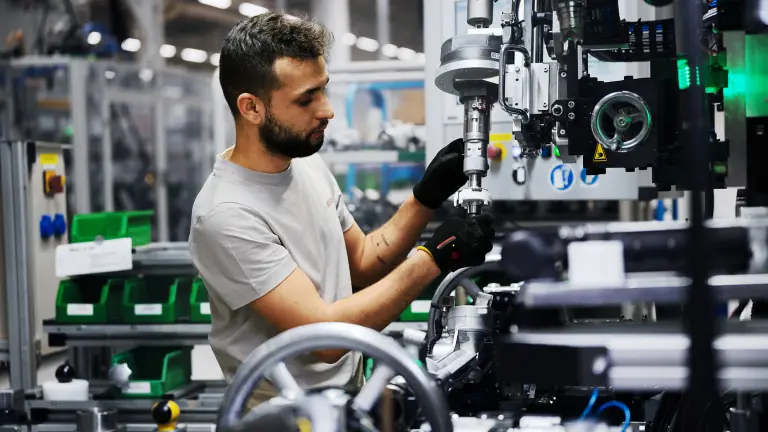 Person with blurred face operates industrial machinery on a factory floor, wearing gloves and handling a mechanical component.