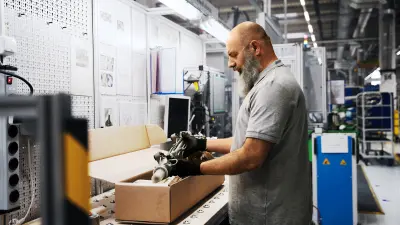 A man stands in a factory in front of a workbench and packs a refurbished steering system into a cardboard box.