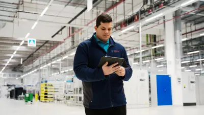 A man is standing in a factory. He has lowered his gaze and is looking at a tablet he is holding in his hands.