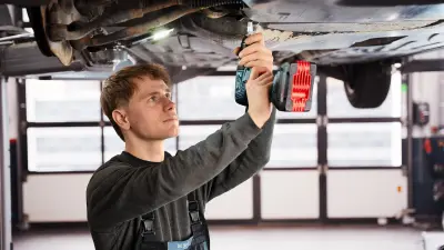 A mechanic is standing under a car that has been lifted on a car lift. He is holding a cordless screwdriver and is working on the underside of the car.