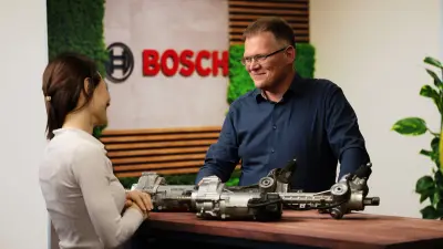 A man stands diagonally opposite a woman in front of the Bosch logo. They smile at each other. A table stands between them. On the table lie an old steering system and a new steering system.