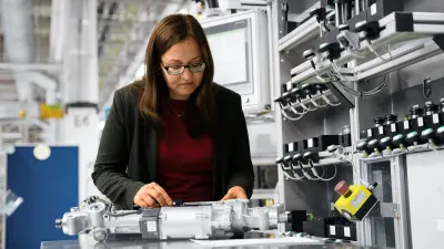 A woman stands in a factory in front of a workbench, checking a steering system for wear. 