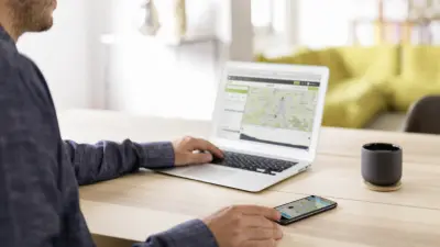 A man sits in a living room at a table and plans his next eBike tour with the help of an app. With one hand he uses the laptop and with the other hand he uses the smartphone.