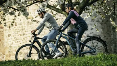 A man and a woman ride eBikes on a path between a meadow and a tree.