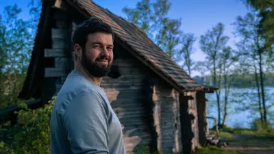 Adam Detre walks toward an old wooden sauna near a lake. He looks back and smiles at the camera.