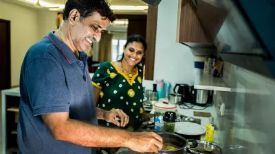 A man with dark, curly hair and a mustache, wearing a blue polo shirt with a red trim, is smiling and looking down while stirring something in a pot on a stovetop in a brightly lit kitchen. To his right and slightly behind him, a woman with dark hair pulled back, wearing glasses and a green patterned dress, is also smiling and looking at him.