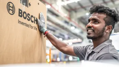A smiling man in a grey t-shirt and gloves stands next to a large brown cardboard box with the black Bosch logo and "Invented for life" text in a factory hall.