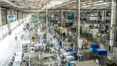 A wide-angle shot shows the interior of a large, brightly lit factory hall with numerous workstations, machinery, and workers involved in appliance manufacturing.