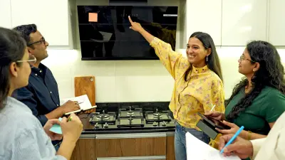 A group of five people, four women and one man, are gathered in a modern kitchen. A smiling woman with long dark hair, wearing a yellow floral blouse and jeans, points upward to a sleek black kitchen hood with integrated lights and controls. She is explaining something about the appliance.
