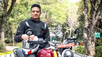 Bosch associate Sandeep C D, a man in a black and grey riding jacket and black pants, sits on a red Triumph motorcycle, holding a white and red helmet. He looks directly at the viewer. The motorcycle is parked on an asphalt road with trees and greenery in the background.