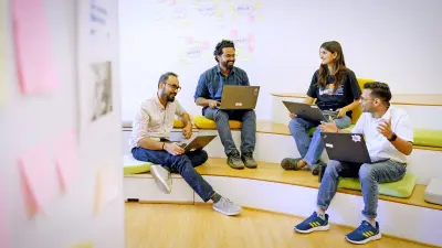 Four people are sitting in a semi-circle on tiered white and light green benches, looking at and using their laptops while smiling and conversing. Two men are on the left, and a woman and a man are on the right.