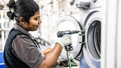 A female factory worker is assembling the door of a washing machine using an electric screwdriver. She is wearing grey gloves and an apron, concentrating on her task.