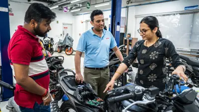 A group of three engineers, two men and one woman, are gathered around a motorcycle inside a workshop. The woman, wearing glasses and a patterned dark top, is pointing at an open compartment on the motorcycle, revealing internal components and circuitry.