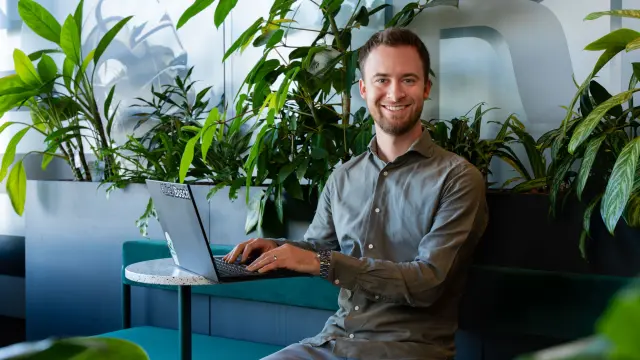 Patrick sits in front of his laptop between houseplants and smiles at the camera. 1