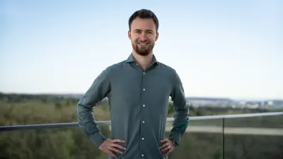 Patrick wears a grey shirt, stands outside in front of the blue sky and smiles at the camera.