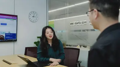 Yunge is sitting on a black chair at a large wooden table in an office environment. In front of her is a black, opened laptop. Her hands are on the table. On the wall in the background is a clock showing about three o'clock and a TV screen showing a Bosch website. Her view is directed towards a man in a black shirt and glasses, who can be seen cropped in the foreground and off from the side.