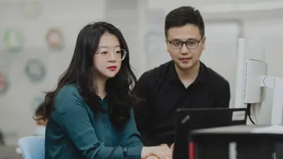 Yunge is sitting at a desk with her hands clasped on the table. Her view is directed at a laptop. Next to it is a large screen. A man in a black shirt and black glasses sits to Yunge's right at the desk.