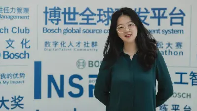 Yunge has her arms crossed behind her back. She is standing in front of a white wall on which various Chinese words can be seen written in blue. She wears glasses and smiles at the camera.
