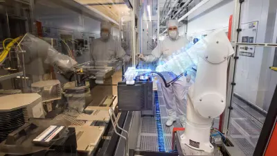 A robot stands in a clean room of a semiconductor manufacturing facility and loads a machine.