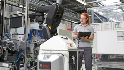 A woman stands with a clipboard and a pen in her hand next to an industrial robot.