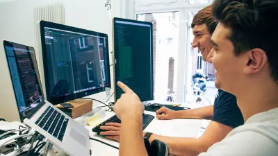 Two young men sit in front of computer monitors and write software codes.