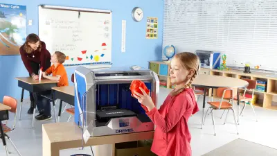 A young girl stands next to a 3d printer in a classroom. She holds something she has printed in her hands.