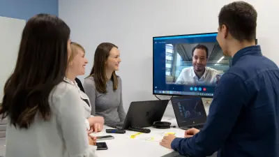 Four people are in a conference room and have a video conference with a fifth person which is on a screen.