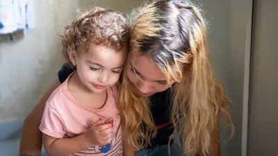 A young woman with long blond hair hugs a child from the side.