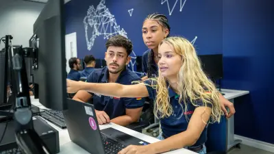 Young woman with blond hair sits at desk and points to the screen. A man sits next to her, a woman stands bent behind both.