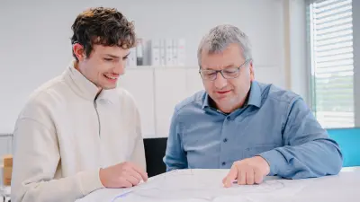 Jonas und sein Teamleiter stehen nebeneinander und blicken beide auf einen Plan, der vor ihnen auf dem Tisch liegt.