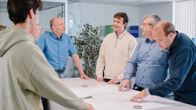 Jonas steht gemeinsam mit seinen Kollegen an einem Tisch für eine Besprechung.