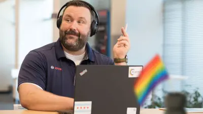 Alfons Müller sitzt vor seinem Laptop. Er trägt ein Headset und hält einen Stift in die Höhe. Es scheint, als hätte er gerade eine gute Idee. Im Vordergrund weht eine kleine Regenbogenfahne.
