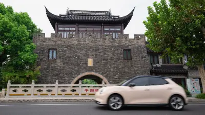 Ora Good Cat drives past the camera on a street in Suzhou. Behind it an old gate to a park.