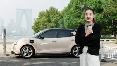 Cao Mengyi steht mit einem Kaffeebecher in Suzhou vor ihrem Auto Ora Good Cat und schaut in die Kamera. Hinter ihr sieht man Bäume eines Parks und Wolkenkratzer.